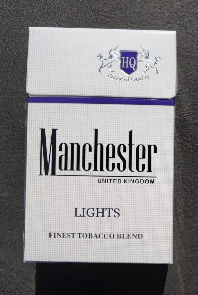 Front of a Manchester Light Blue cigarette pack photographed on a black textured surface